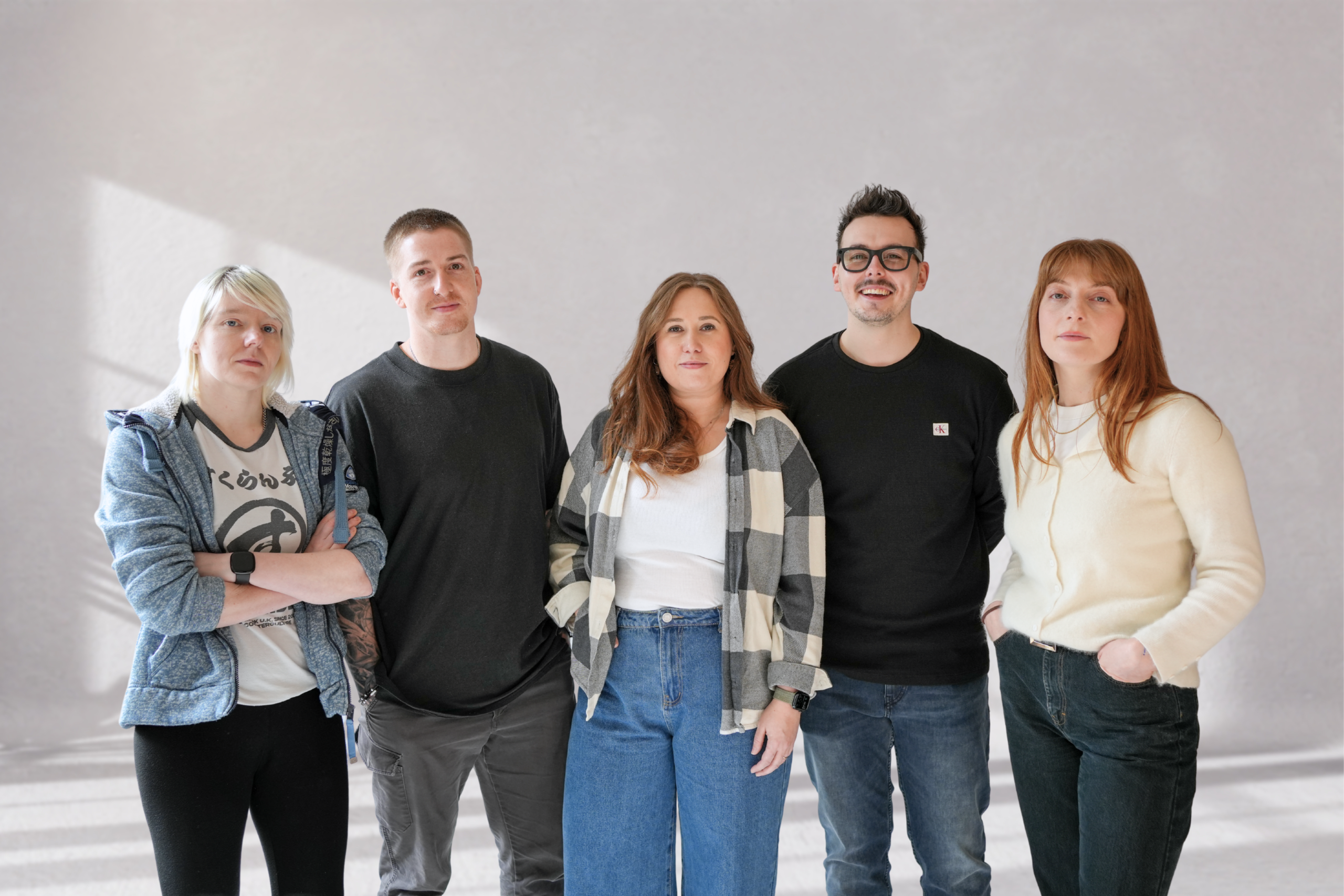 A group photograph of the Search Theory team, which visualises five team members. Two male and two female. In the photo the team are standing in a photograph studio and are smiling towards the camera, like a cool indie band.